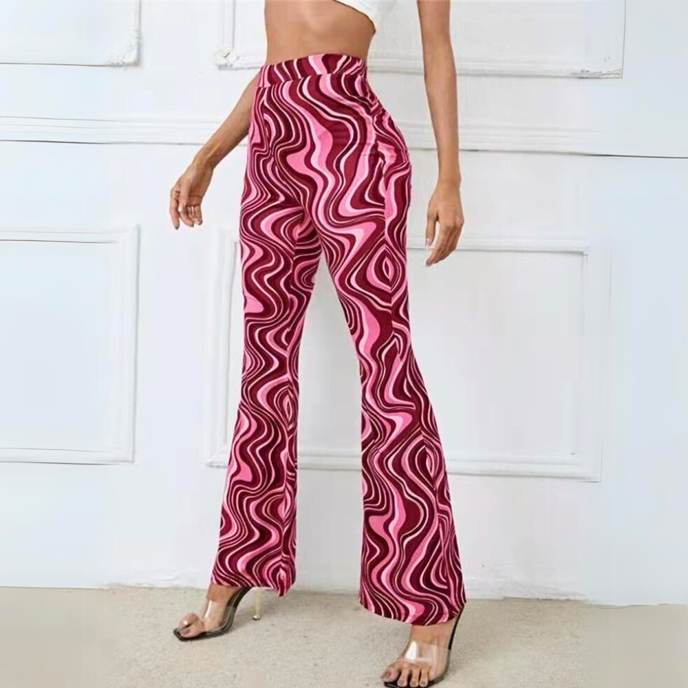 A woman stands indoors wearing trendy statement pants—high-waisted, wide-leg trousers with a vibrant pink and maroon wavy geometric pattern—paired with clear high-heeled sandals and a white crop top.