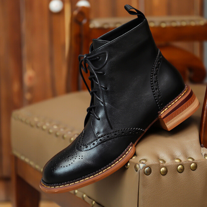 A black leather brogue boot with lace-up design and brown sole is displayed on a tan, gold-studded upholstered chair in a warmly lit room—perfect for the urban trendsetter seeking genuine leather style.