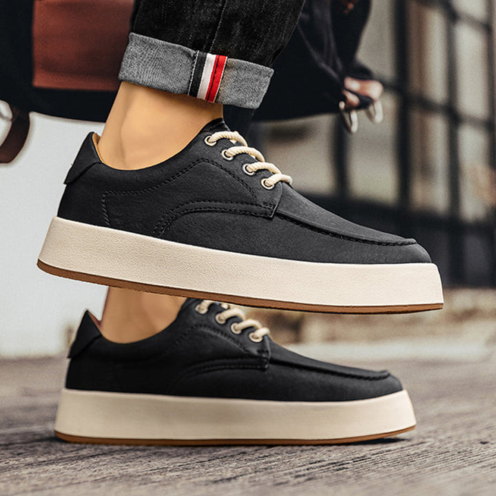 A person wearing black men's leather casual shoes with white soles and beige laces stands on a wooden surface. The jeans are slightly rolled up, revealing a red and white stripe on the cuff for a touch of Italian designer style.