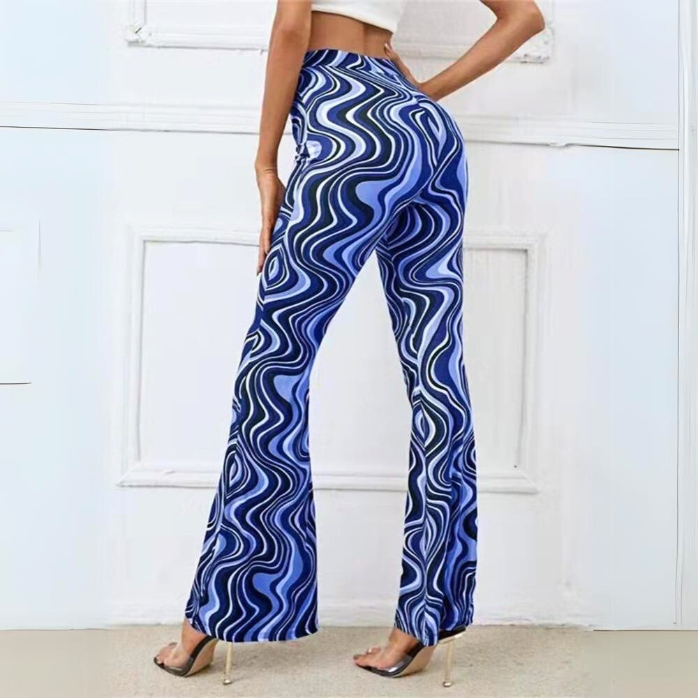 A person wearing a white crop top and trendy statement pants—high-waisted, flared with a blue and white wavy pattern—stands with one hand on their hip against a white wall, modeling clear high-heeled shoes.