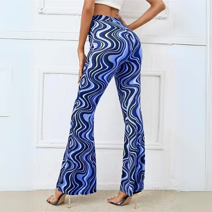 A person wearing a white crop top and trendy statement pants—high-waisted, flared with a blue and white wavy pattern—stands with one hand on their hip against a white wall, modeling clear high-heeled shoes.