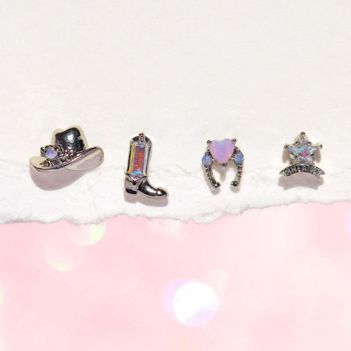 Four small, sparkling statement earrings on torn white paper: a cowboy hat, boot, horseshoe with a heart, and sheriff badge, all with iridescent or gem-like details, bring urban chic flair against a soft pink background.