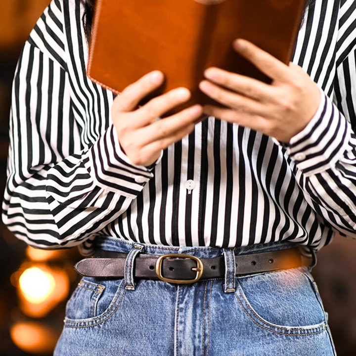 A person wearing a black-and-white striped shirt and blue jeans with a leather belt is holding and reading an open book, seen from the chest down.
