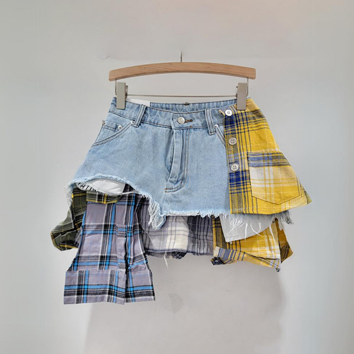 A denim and plaid patchwork skirt featuring layered, colorful plaid fabric in yellow, purple, and blue drapes from the waistband, capturing eclectic fashion vibes on a wooden hanger against a plain background.