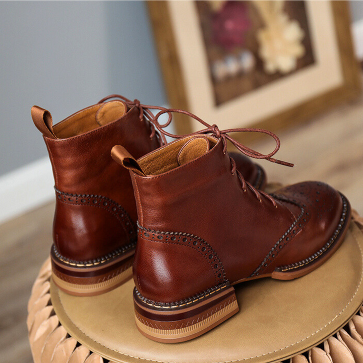 A pair of brown leather brogue boots with decorative perforations, crafted from genuine leather, placed on a woven surface with a blurred framed picture in the background.