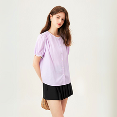 A woman with long brown hair wears a light purple versatile blouse with short puff sleeves and a black pleated skirt, standing against a plain light background, holding a small woven bag behind her back.