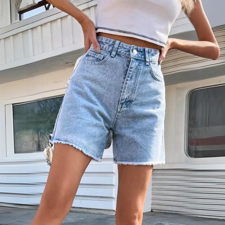 Woman wearing a white crop top and high-waist denim shorts stands outdoors with one hand on her hip. Only her torso and legs are visible, showcasing a classic streetwear shorts look.