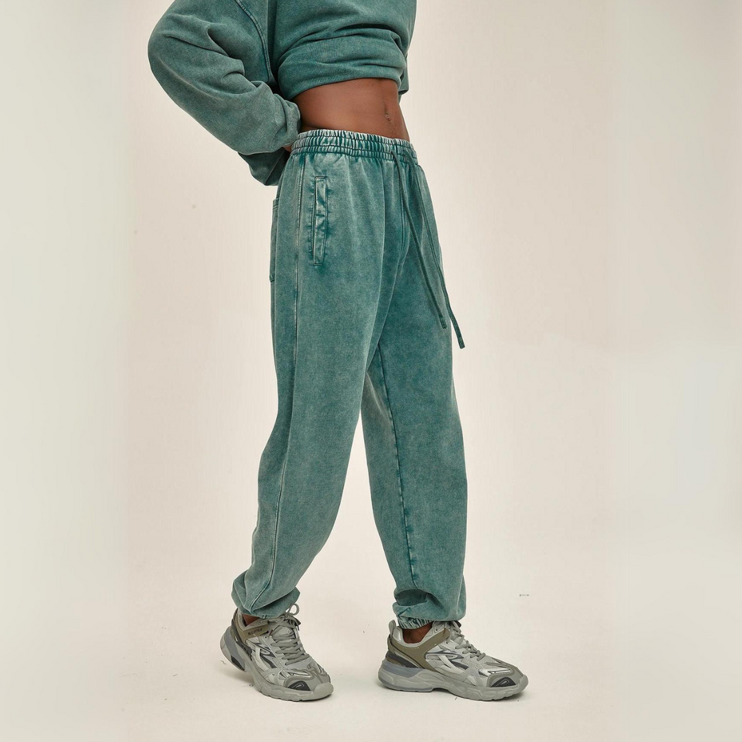 A person wearing a green cropped sweatshirt and matching loose jogger pants with an elastic waistband and tapered cuffs, paired with silver-gray sneakers, stands against a plain off-white background. Their face is not visible.