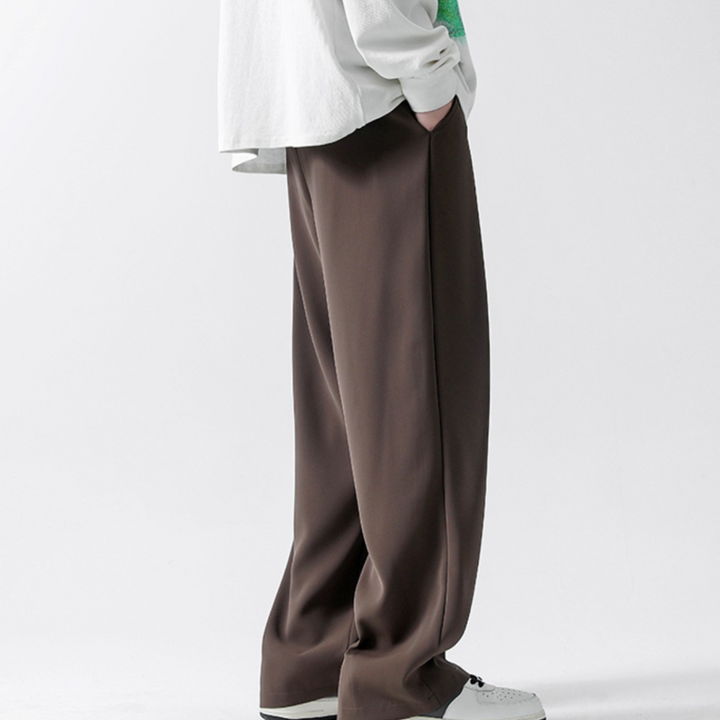 Person standing with hands in pockets, wearing loose-fitting brown streetwear joggers, a white long-sleeved shirt with green details, and white sneakers. The background is plain and light-colored.