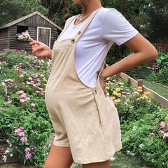 A pregnant person in comfortable maternity clothes—a pair of beige pregnancy overalls and a white T-shirt—stands in a colorful flower garden, holding a bunch of flowers with one hand on their hip. A wooden shed sits in the background.