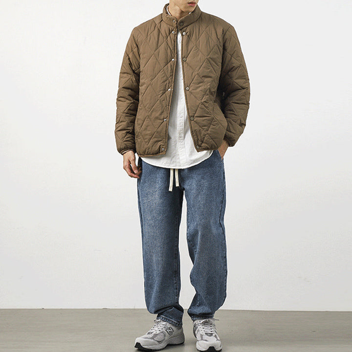 A person stands against a plain white background wearing a brown lightweight insulating padded jacket with a diamond-stitched pattern, a white shirt, loose blue jeans, and gray sneakers. Their hands are in their pockets and their face is not fully visible.