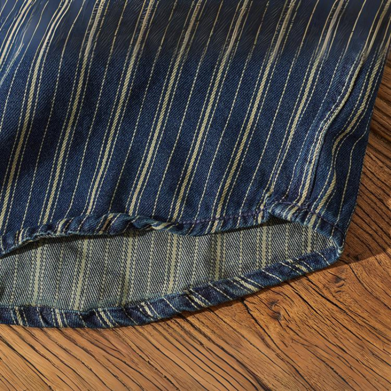 A close-up view of a striped denim shirt with vertical white pinstripes, draped over a wooden surface. The fabric edge is visible with detailed stitching along the hem.