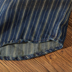 A close-up view of a striped denim shirt with vertical white pinstripes, draped over a wooden surface. The fabric edge is visible with detailed stitching along the hem.