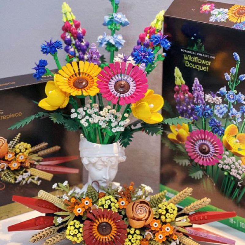 Colorful LEGO Wildflower Bouquet and floral designs are displayed in front of their packaging. The flowers, built from building blocks, are arranged in a white vase shaped like a classical statue’s head.