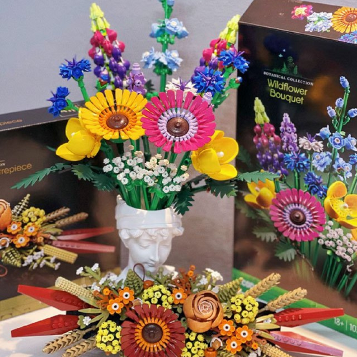 Colorful LEGO Wildflower Bouquet and floral designs are displayed in front of their packaging. The flowers, built from building blocks, are arranged in a white vase shaped like a classical statue’s head.
