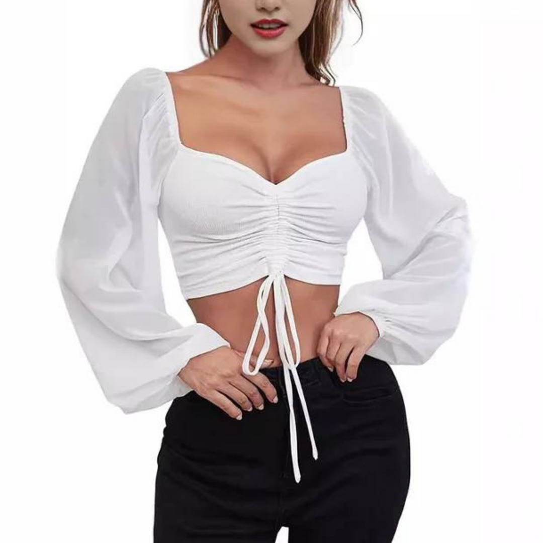 Woman wearing a white, long-sleeve crop top with a ruched front and drawstring, featuring a flirty neckline, paired with black high-waisted pants and standing with one hand on her hip.