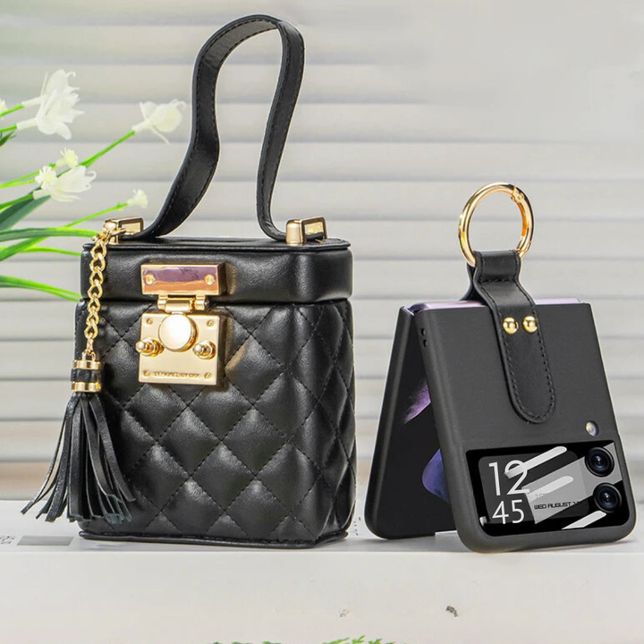 A small, black, quilted handbag with gold hardware and a tassel sits beside a leather phone case with a gold built-in ring holder, holding a folded phone displaying the time 12:45. Flowers are blurred in the background.
