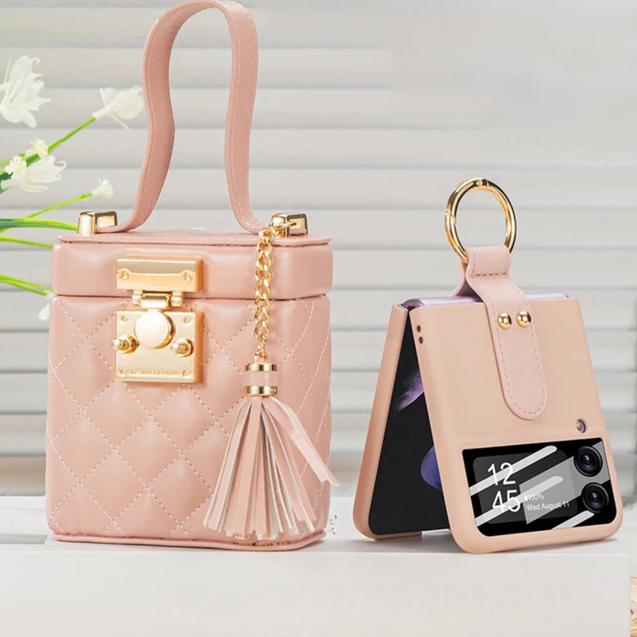 A small pink quilted handbag with a gold lock, chain, and tassel sits next to a matching leather phone case holding a folded smartphone with a built-in ring holder. A keyring is attached, and white flowers are visible in the background.