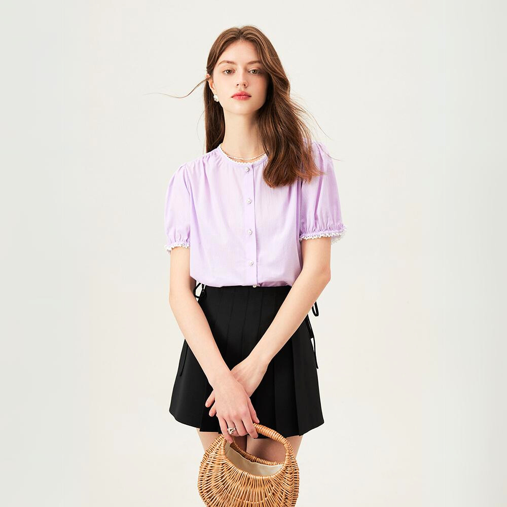 A young woman with long brown hair stands against a plain background, wearing a versatile light purple button-up blouse and a black pleated skirt. She holds a round wicker handbag in front of her with both hands.