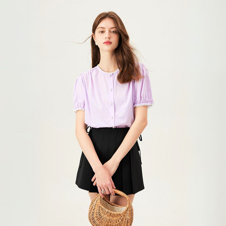 A young woman with long brown hair stands against a plain background, wearing a versatile light purple button-up blouse and a black pleated skirt. She holds a round wicker handbag in front of her with both hands.