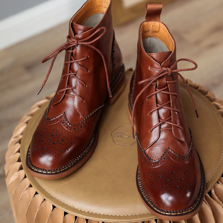 A pair of polished brown genuine leather brogue ankle boots with decorative perforations and laces, displayed on a round tan ottoman with a wooden floor in the background—perfect for the urban trendsetter.