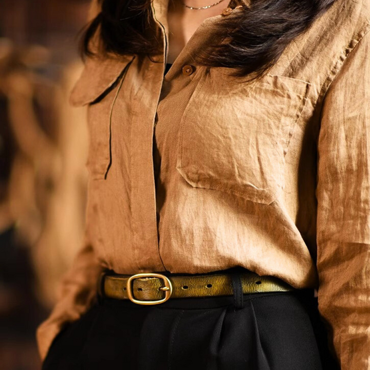 A person wears a light brown button-up shirt tucked into high-waisted black pants, accented with a green leather belt featuring a gold buckle. The image captures classic country fashion, focusing on the torso with the head partially out of frame.