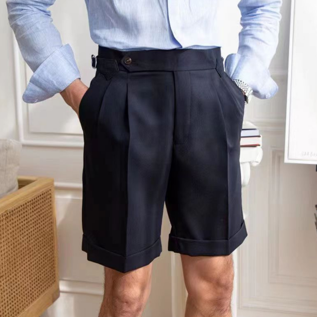 A person wearing a light blue dress shirt tucked into high-waisted pleated shorts with turn-up hems, showcases the old money aesthetic indoors, hands casually in pockets for a refined yet relaxed tailored fit.