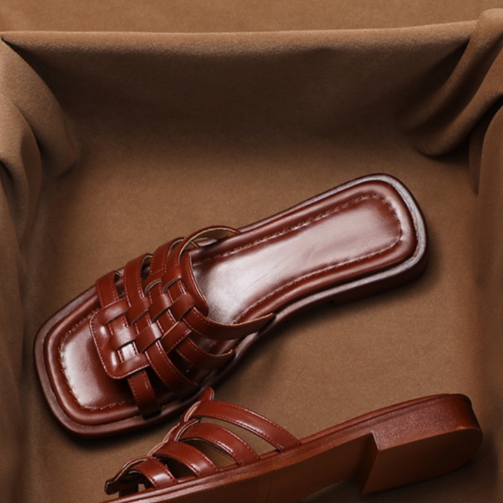 A pair of brown, woven Italian leather slide sandals with square toes and low heels, featuring a comfort cushioned insole, displayed on a soft brown fabric background.