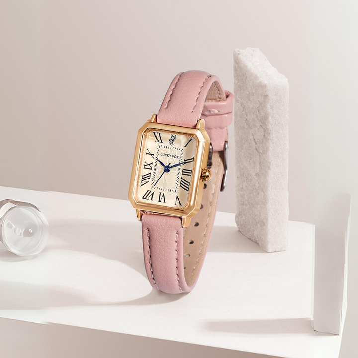 A rectangular face watch with a gold case, white dial, and black Roman numerals sits upright on a white surface. Its genuine leather strap in pink complements the geometric white background props.