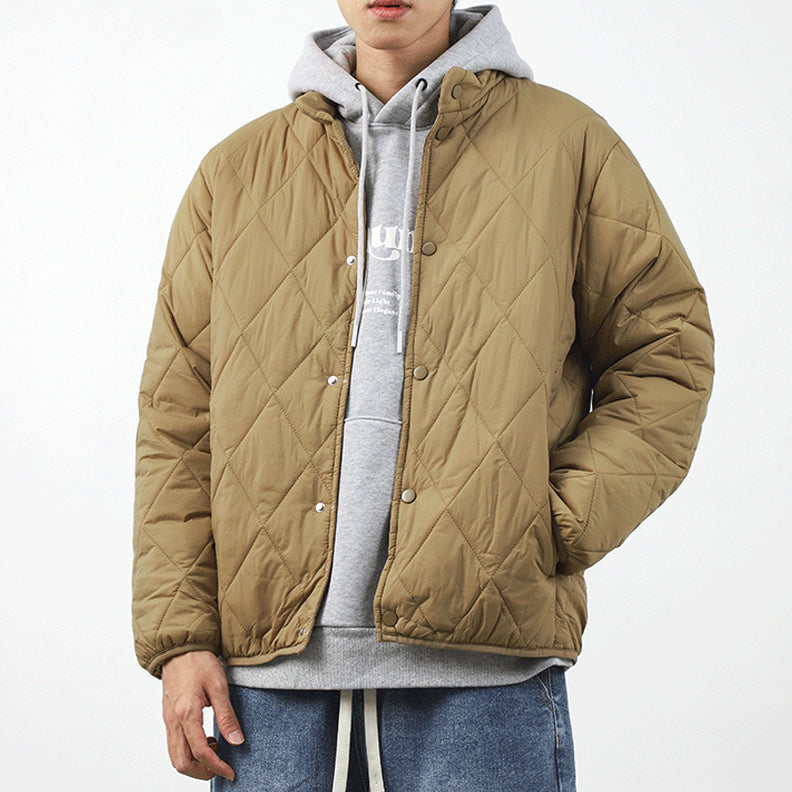 A person wearing a light brown padded jacket with a diamond-stitched pattern over a gray hoodie and blue jeans stands against a plain white background. The lightweight insulating jacket is buttoned up, and the persons head is partially out of frame.
