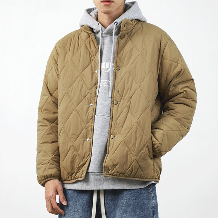 A person wearing a light brown padded jacket with a diamond-stitched pattern over a gray hoodie and blue jeans stands against a plain white background. The lightweight insulating jacket is buttoned up, and the persons head is partially out of frame.