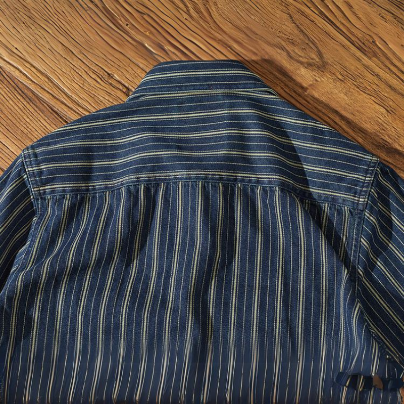 A blue striped denim shirt with relaxed-fit vertical white and yellow stripes is laid flat on a wooden surface, displaying the back and collar.
