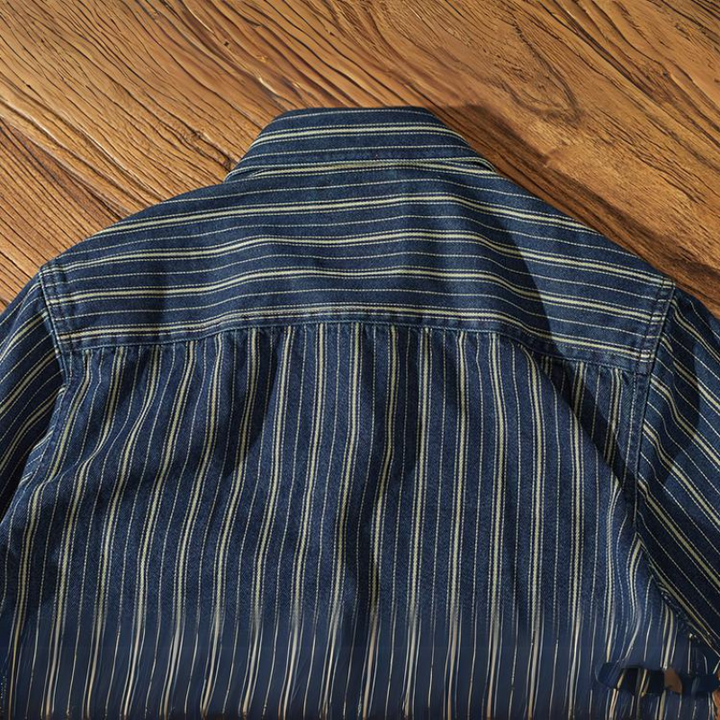 A blue striped denim shirt with relaxed-fit vertical white and yellow stripes is laid flat on a wooden surface, displaying the back and collar.