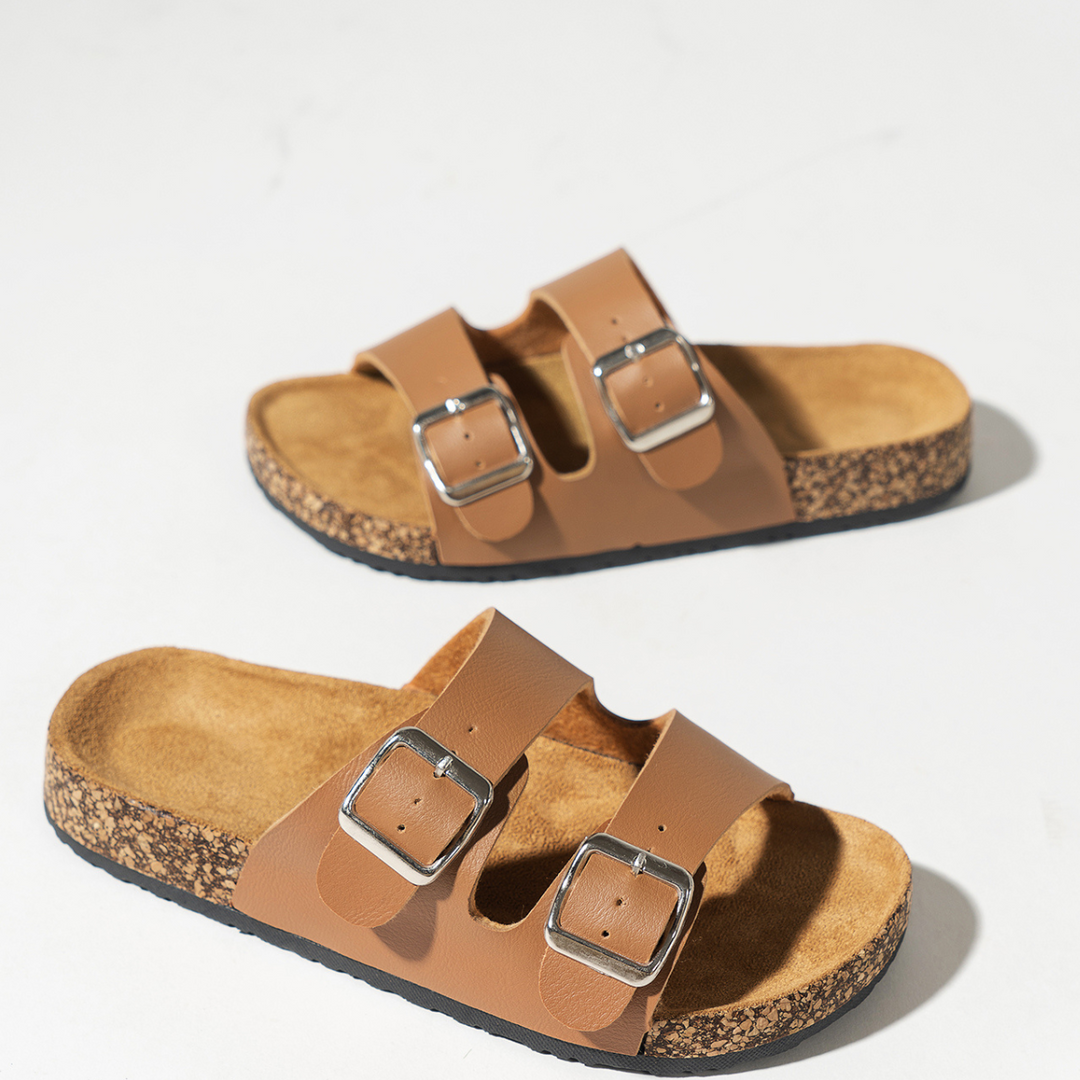 A pair of tan double-buckle sandals with premium leather straps and cork-like soles are placed on a white surface.