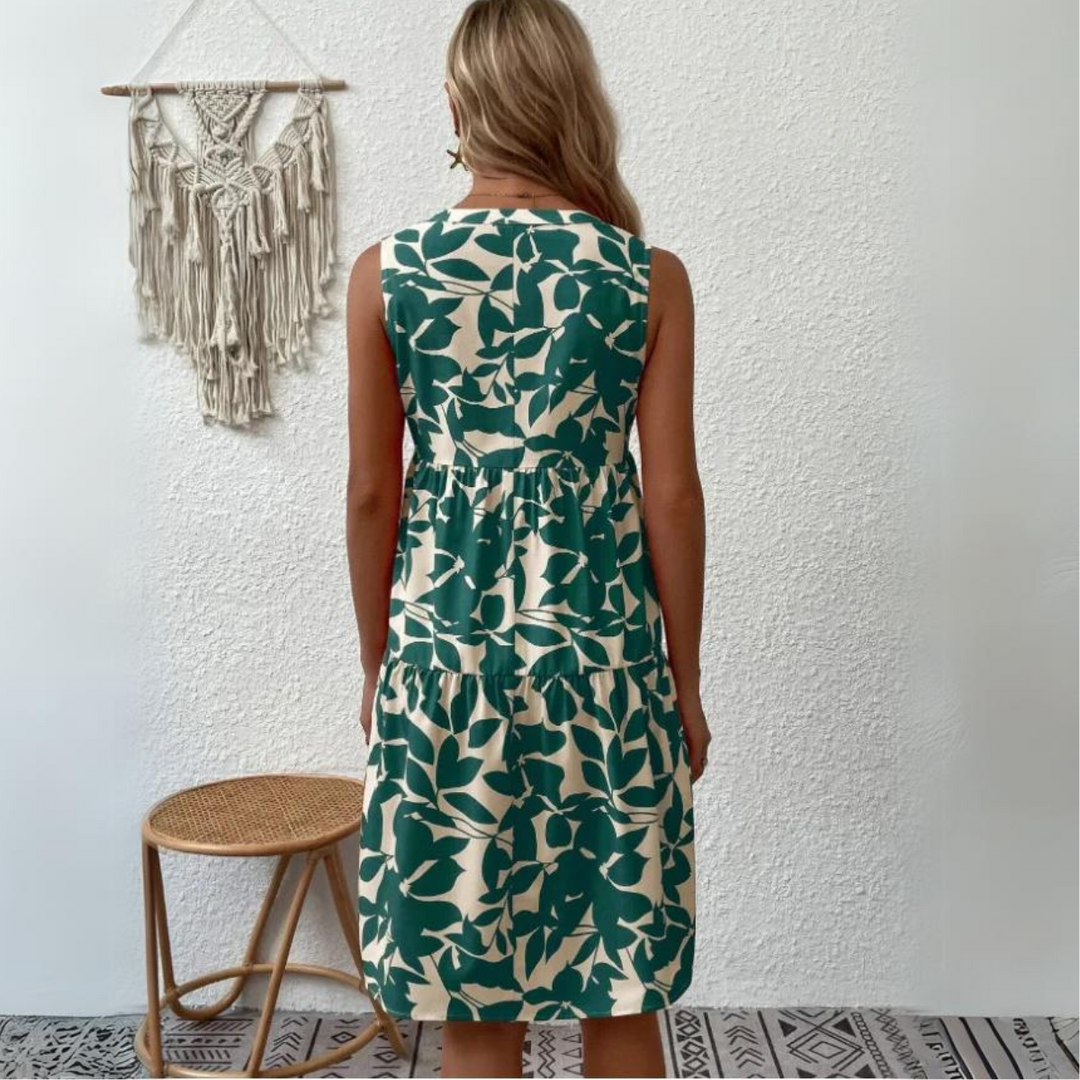 A woman with blonde hair, wearing a sleeveless dress crafted from lightweight breathable fabric with green leaf patterns, stands facing a white textured wall. A wooden stool and a macramé wall hanging are on her left. Perfect for your summer wardrobe.