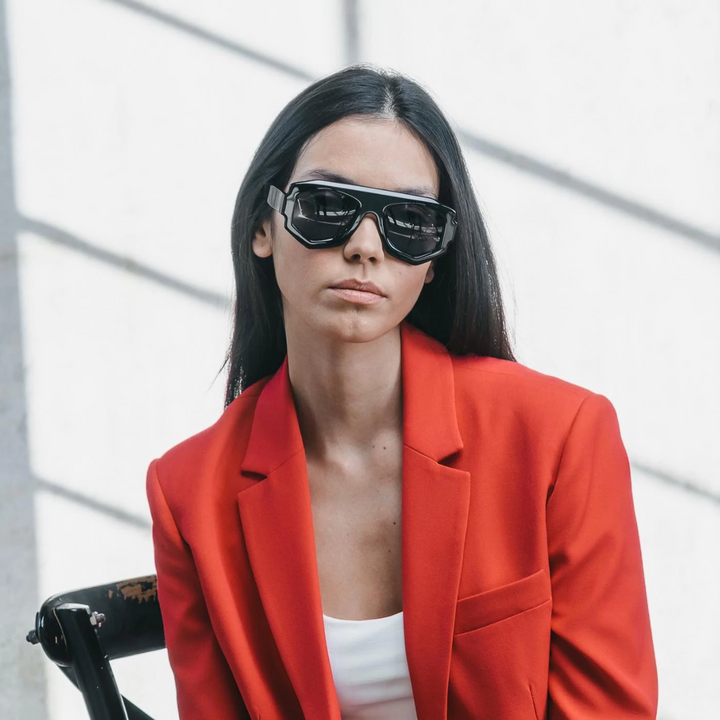 A woman with straight dark hair wears oversized black geometric frame sunglasses and a bright red blazer over a white top, sitting on a chair against a white wall with window shadows.
