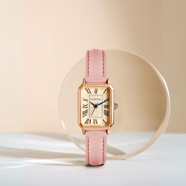 A gold rectangular face watch with a genuine leather strap in pink is displayed upright against a clear oval prop on a beige background. The Roman numeral watch features blue hands for an elegant touch.
