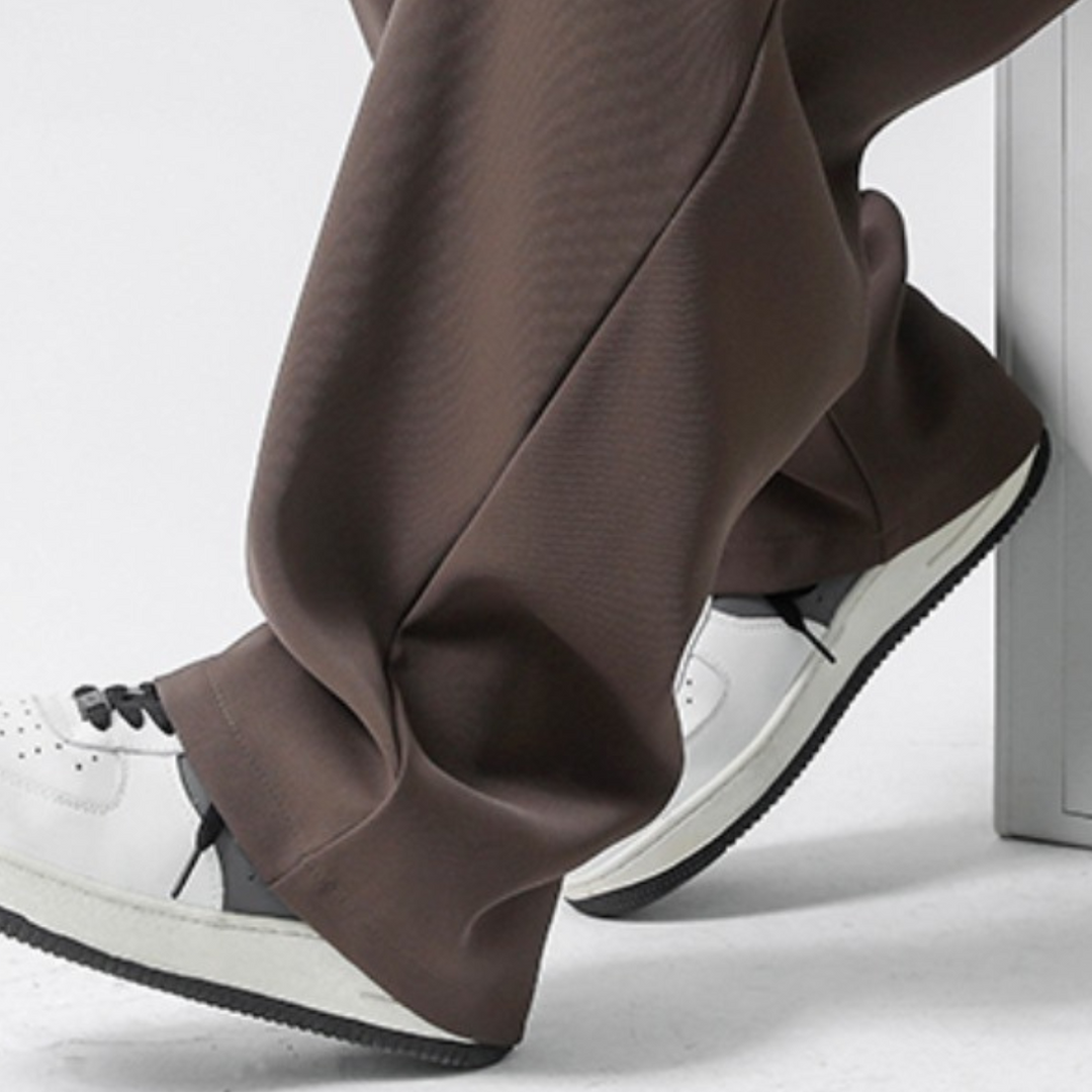 Person wearing loose brown streetwear joggers and white sneakers with black details, sitting with one foot on the floor and the other on a low surface. The photo is cropped to show only the lower legs and feet.