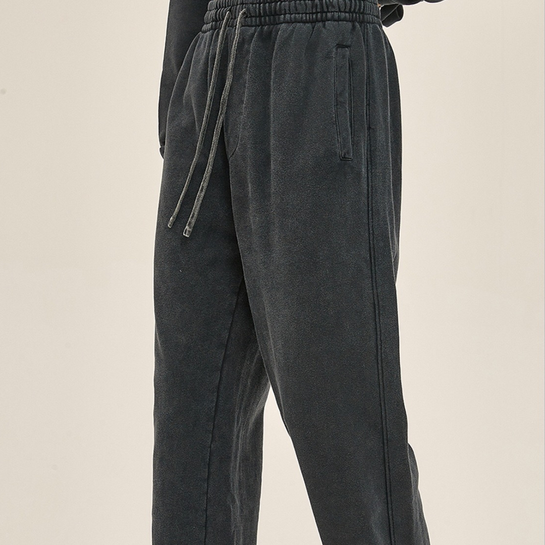 Person wearing loose-fitting, dark gray sweatpants with an elastic waistband and deep pockets, standing against a plain light background. Only the lower half of the body is visible.