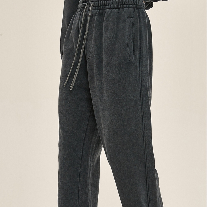 Person wearing loose-fitting, dark gray sweatpants with an elastic waistband and deep pockets, standing against a plain light background. Only the lower half of the body is visible.