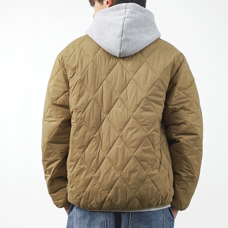 A person wearing a tan, lightweight insulating, diamond-stitched jacket over a gray hooded sweatshirt, seen from behind. The individual has short hair and is also wearing blue jeans. The background is plain and white.
