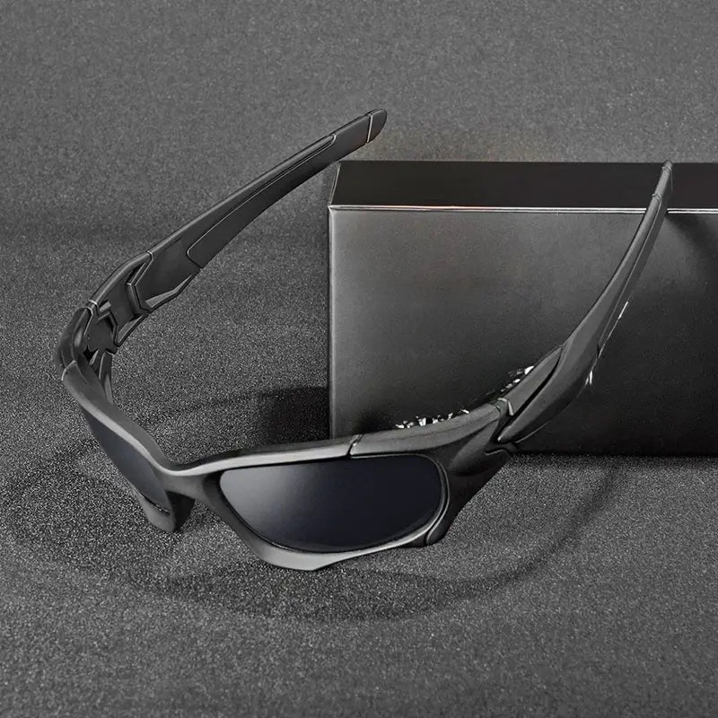 Sleek black wraparound sports sunglasses with dark, interchangeable lenses are displayed on a textured surface next to a black rectangular box.