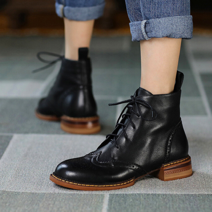 A person wearing cuffed blue jeans and genuine leather black lace-up brogue boots with brown soles is standing on a tiled floor, embodying the style of an urban trendsetter.