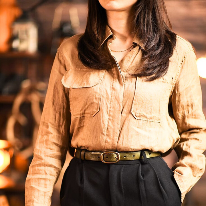 A woman wearing a light brown button-up shirt tucked into high-waisted black pants with a leather belt, standing indoors against a blurred, warmly lit background. Her face is partially out of the frame, channeling effortless country fashion.