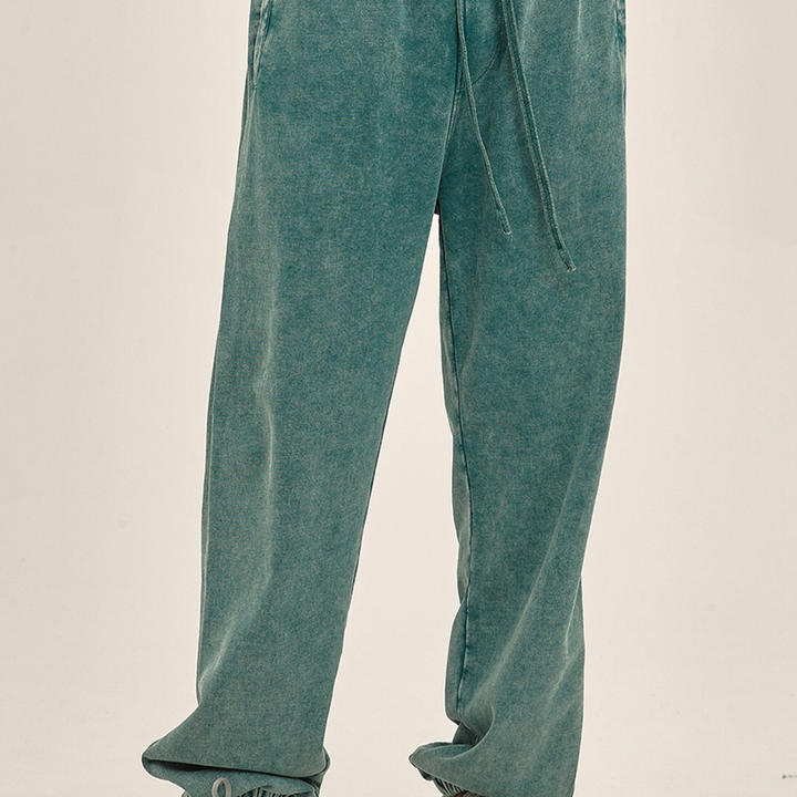 Person wearing loose-fitting, teal-colored sweatpants with an elastic waistband and tapered cuffs, standing against a plain, light background. Only the lower half of the body is visible.