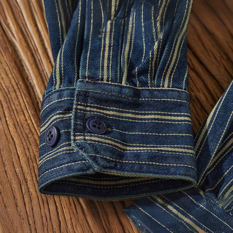 A close-up of a relaxed-fit button-down striped denim shirt sleeve with vertical tan stripes, featuring a cuff with two dark blue buttons, resting on a wooden surface.