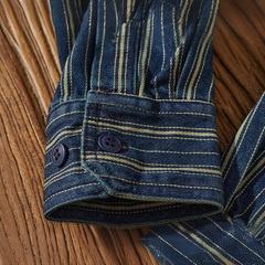 A close-up of a relaxed-fit button-down striped denim shirt sleeve with vertical tan stripes, featuring a cuff with two dark blue buttons, resting on a wooden surface.