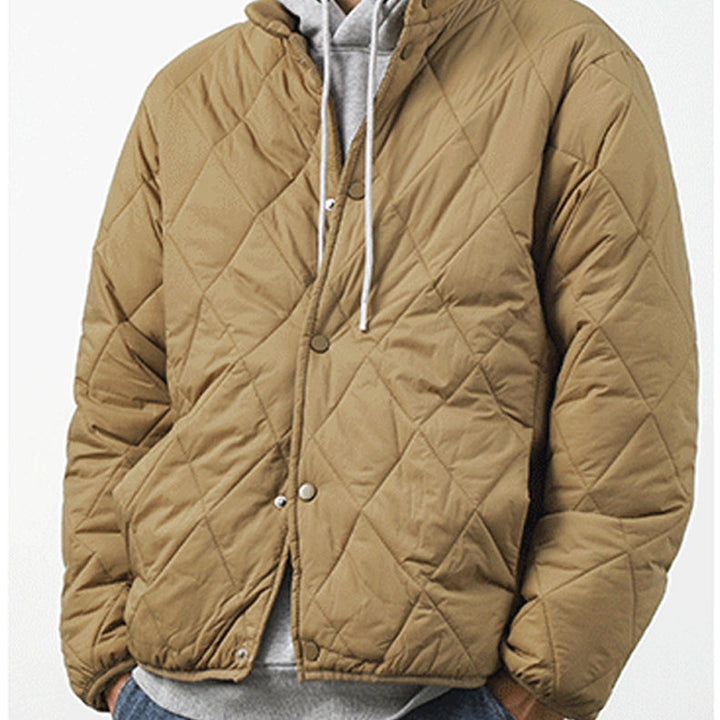 A person wearing a tan, diamond-stitched padded jacket over a gray hoodie with white drawstrings, standing against a plain light background.
