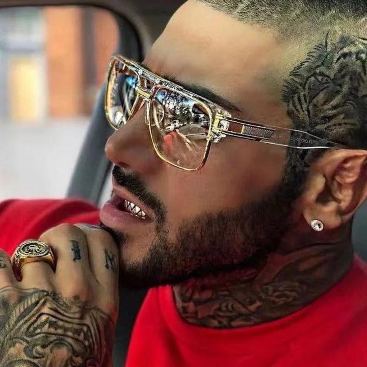 A man with tattoos, a close-cropped haircut, and a beard wears large geometric sunglasses with UV400 protection, a diamond stud earring, and a red shirt. He has a gold ring and is sitting in a car.