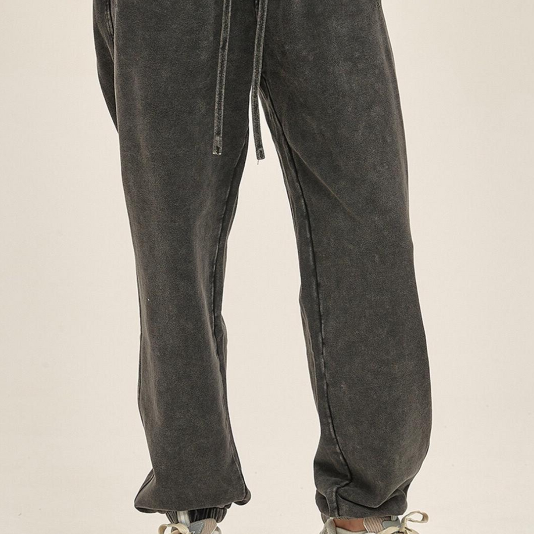 A person wearing loose-fitting, faded black jogger pants with an elastic waistband and drawstring, paired with light-colored sneakers. The photo is cropped to show only the lower half of the body.